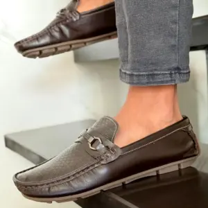 Mocasines de conducción en piel genuina café oscuro con empeine texturizado, antifaz metálico y suela segmentada en caucho, fotografiados en un escalón.